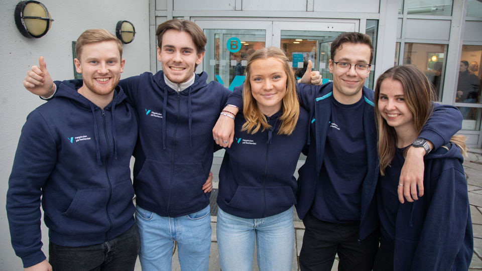 Smilande studentar oppstilt på rekke. Foran inngangen til campus Stord.