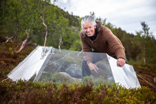 Stein Joar Hegland står bøygd over eit drivhus med open topp og smiler. Han har halvlangt hår og er kledd i mosegrøne bukser og brun genser. Han er omringa av tre og blåbærlyng.