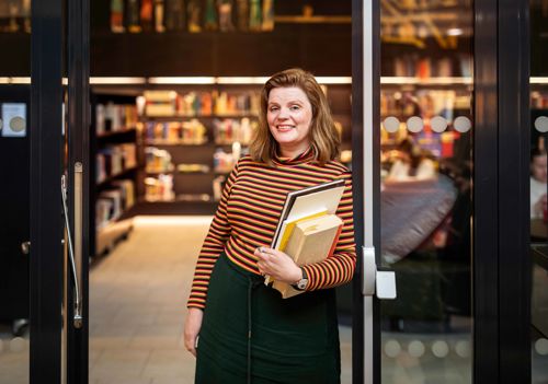 Professor Edit Bugge står i døra til biblioteket på campus Bergen. Ho smiler. Edit har raudleg hår og er kledd i ein genser med svarte og oransje striper og eit grønt skjørt. Ho held eit utval bøker av ulik storleik.