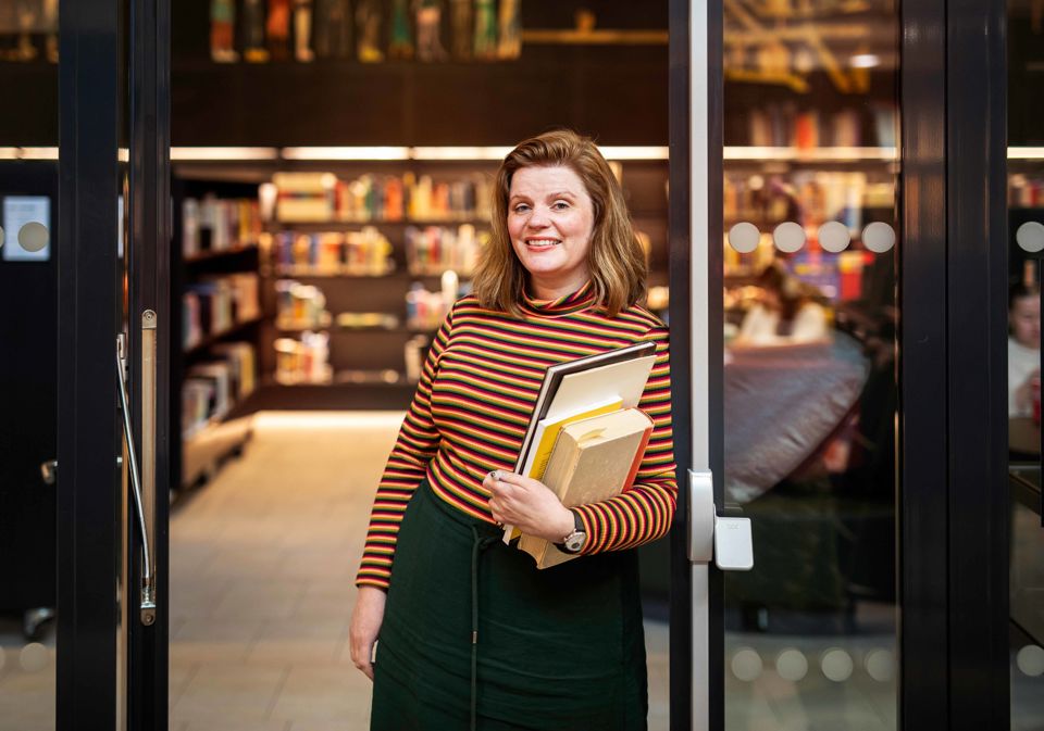 Professor Edit Bugge står i døra til biblioteket på campus Bergen. Ho smiler. Edit har raudleg hår og er kledd i ein genser med svarte og oransje striper og eit grønt skjørt. Ho held eit utval bøker av ulik storleik.
