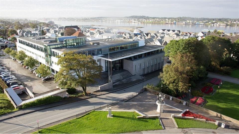 En illustrasjon av nybygget i Haugesund. Illustrasjonen viser et stort og moderne bygg med mange vinduer og grønne parkområder på siden. Bak i bildet kan man se sjøen.