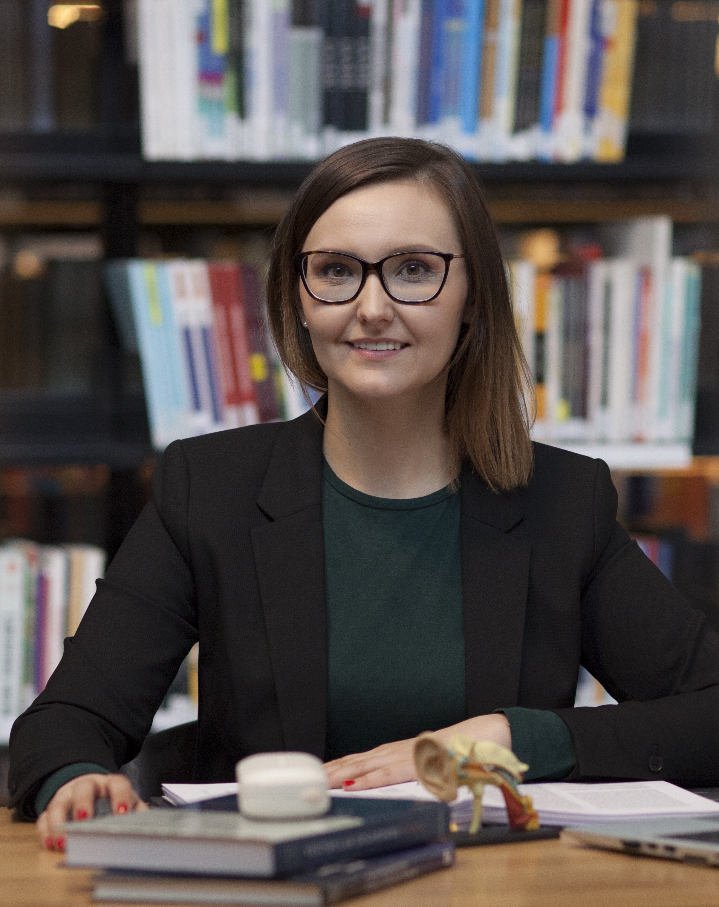 Sylwia Kolasa, en person med rett brunt hår og briller, iført en grønn skjorte med en svart blazer over. Hun sitter bak et slags bord med diverse bøker og greier på. I bakgrunnen ser vi ei bokhylle med mange bøker med fargerike permer.